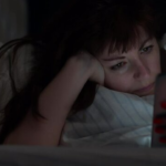 A tired woman lying in a messy bed looking at her phone in a dark room.