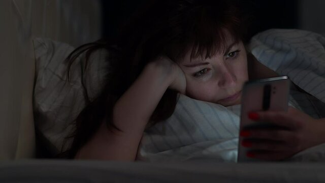 A tired woman lying in a messy bed looking at her phone in a dark room.