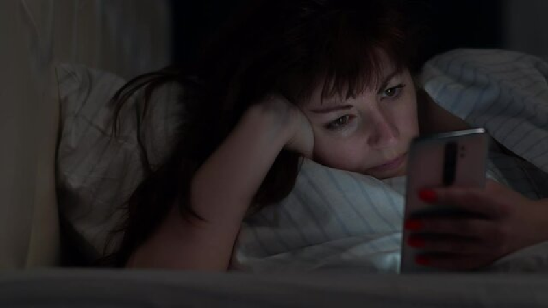 A tired woman lying in a messy bed looking at her phone in a dark room.